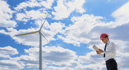 Young man engineer in white shirt and red safety helmet holding a tablet and verifying the wind turbine windmill.