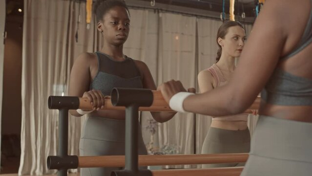 Young Black woman leaning on gymnastic barre in fitness studio doing squats on one leg in front of mirror with other women