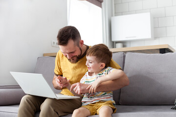 Father teaching his little son to use laptop at home. Family relax on a couch, funny study, learning computer, working, spending time together. Selective focus.