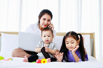 little daughter and Baby play baby toys together on bed while Busy mother working on laptop