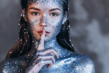 Young beautiful girl in fantastic creative makeup with rhinestone presses finger to mouth, dark background. Art woman model with cosmic make-up on face and body blue and silver skin color 