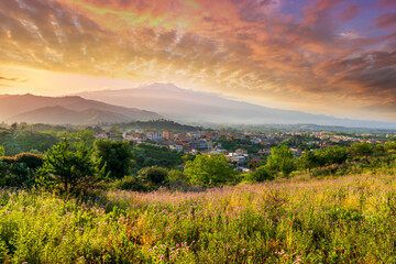 view from hill with golden grass and green bushes to a valley town with majectic mountains and scenic cloudy sunset on background