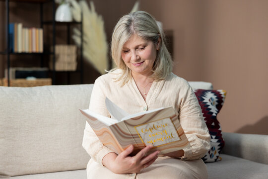 Middle-aged woman relaxing on couch looking at photo album she found while cleaning, reminiscing about her youth, wedding, laughing when she sees pictures of daughter son misses being alone.