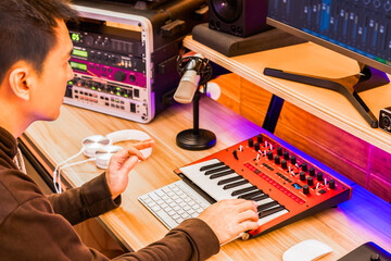 asian professional music producer playing midi keyboard synthesizer for arranging a hit song on computer in home studio showing recording equipments for music production