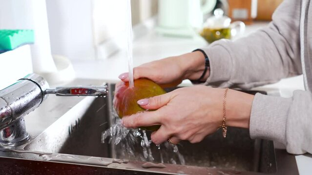 A woman washes a ripe and juicy mango under a stream of water in the kitchen.