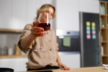 Selective focus of lonely woman in kitchen making toast while raising glass of red wine. Mature female celebrating something