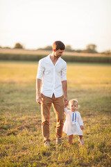 Fototapeta premium Beautiful young dad and his little blue-eyed curly 1 year old daughter in light natural clothes are walking on the sunset in the summer in the field. Papa's love. Family. Sunset.