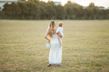 Naklejka na ściany i meble Young beautiful blonde curly-haired mother with her little blue-eyed baby girl is walking at sunset in a field. Warm summer evening. Freedom. Sunset.