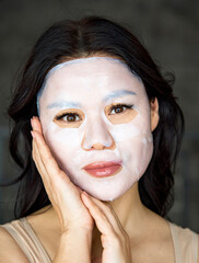 Sensual photo of beautiful young asian smiling woman, applying cotton facial moisturizing mask on face, takes care of skin, prevents wrinkles, stands on isolated grey background