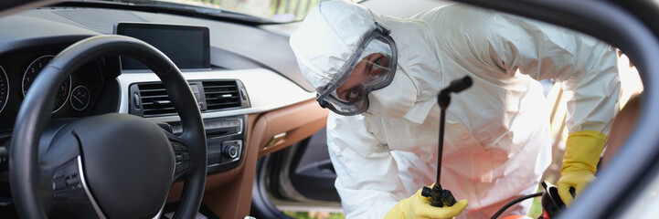 Person in protective costume clean surfaces in car with disinfectant spray