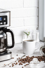 A white cup with a drink on the kitchen table next to a drip coffee maker. Roasted coffee beans. Morning in the kitchen