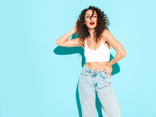 Sexy young beautiful smiling female in trendy summer hipster clothes. carefree woman posing near blue wall in studio. Positive brunette model with curls hairstyle. Cheerful and happy