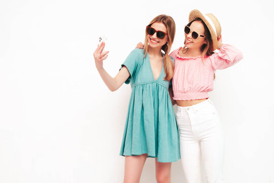 Two young beautiful smiling brunette hipster female in trendy summer dresses. Sexy carefree women posing near white wall. Positive models having fun. Cheerful and happy. In hats. Taking selfie