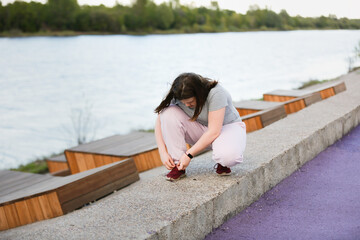 Fototapeta na wymiar Overweight European teenage girl in tracksuit ties her shoelaces on her sneakers while jogging along on concrete embankment, Sports and teenagers, overweight teenagers