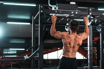 Back view of muscular sportsman doing pull ups on horizontal bar  at gym. Sports trainer in gym...