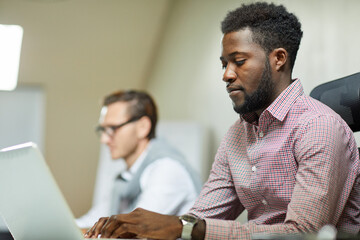 Concentrated young African-American programmer in checkered shirt sitting at table and typing on laptop