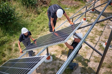 Workers installing solar panel on metal beams in field at sunny daytime. Renewable and ecological energy. Environment safe. Modern technology and innovation. European man wearing workwear and helmets