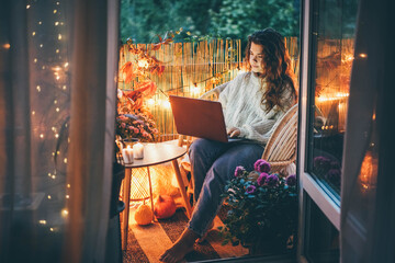 Person sits on chair and types on laptop keyboard put on table near cup and lit candles against wall decorated with lights and autumn attributes in evening