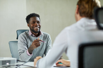 Cheerful young Black recruiter sitting at table and asking questions to job candidate during meeting