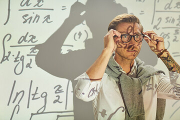 Serious pensive young math analyst with sweater around neck adjusting eyeglasses while preparing for solving equations