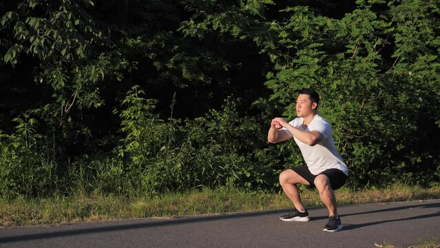 Side view full length shot of active fit young korean man doing squat exercise during workout at street sunset background of green trees. Summer time male workout spot outdoor. Sport healthy concept.