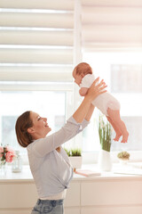 Pretty happy young Caucasian woman in casual clothes lifting her newborn daughter