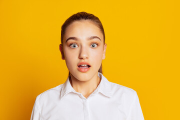 Beautiful surprised girl with long loose hair looking at camera with wide eyes isolated on yellow background. Concept of beauty, art, fashion, facial expression and emotions