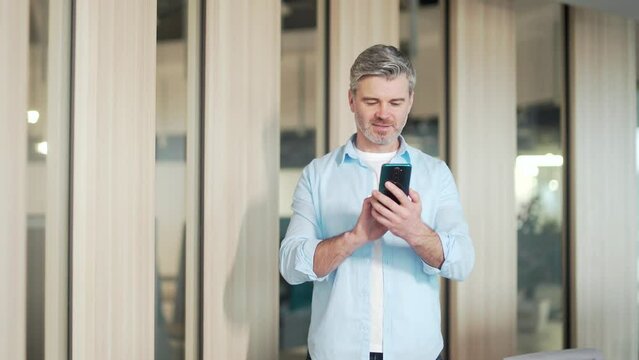 Happy mature gray-haired bearded business man in casual clothes using cellphone standing in modern office. Senior employee browses smartphone. Reads good news smiling. worker chatting looking at smart