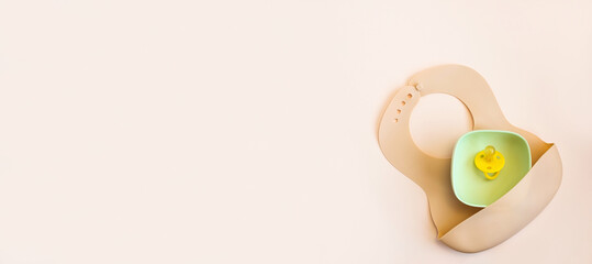 Baby accessories and tableware for food on beige background. First food for kids concept. Flat lay, Top View, Copy Space. Silicone plate, bib with on neutral background. Baby serving