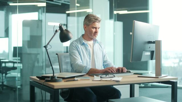 mature gray-haired confident business man in casual clothes working at pc computer workplace in a modern glass office at desk. Focused mature senior male bearded businessman, employee at work