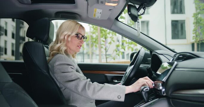 Female mature driver in formal clothes adjusting radio dial button control, gps or air conditioning while sitting behind the whel of car. Inside car view
