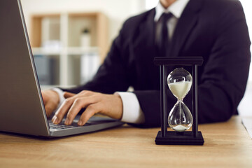 Close up of black hourglass with white trickling sand on table of businessman working on laptop. Hourglass at workplace of man in suit. Business, time and deadline concept. - obrazy, fototapety, plakaty