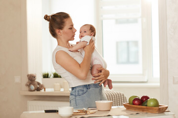 Young mother in white Tshirt talking to her baby while standing in kitchen next to table with food