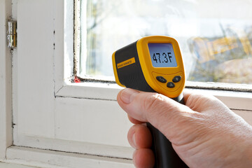 Male hand with an infrared or laser thermometer, measuring the temperature of a window seal where...