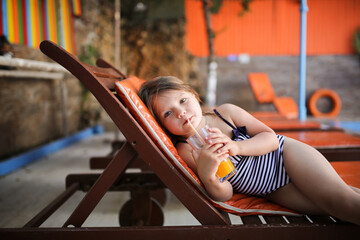 Cute child girl in wide-brimmed hat and sunglasses in swimsuit on wooden sun lounger orange, summer...