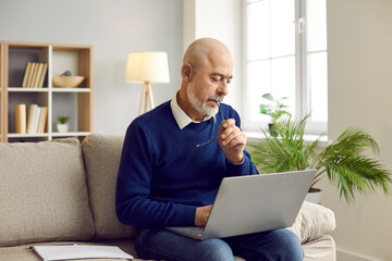 Man using laptop computer at home. Bald bearded senior man sitting on sofa in living room, working...