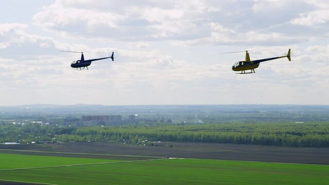 Two bright colored Robinson helicopters fly over green fields against sky with clouds on sunny day. A yellow Robinson helicopter flies behind blue helicopter against summer landscape. 