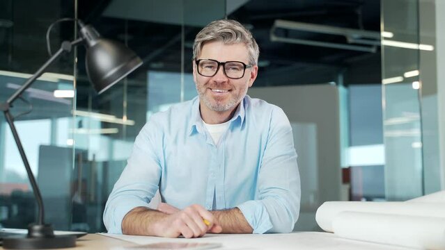 portrait mature bearded gray-haired man creative designer, architect or engineer sitting in office computer desk, looking at camera. Happy senior male employee in glasses on workplace, smiling. indoor