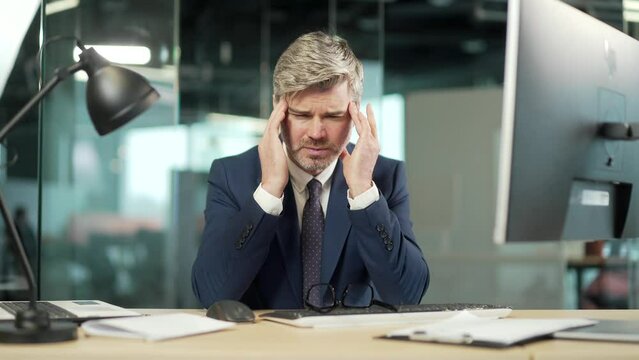 Exhausted, overworked mature office worker sitting in workplace with a severe headache massaging head. unhealthy Tired business male employee in pain ache sick Stress at work sedentary at computer