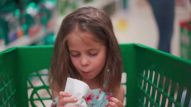 Child sitting shopping cart in store store eats tasty roll churras. Quick snack for children. Backing snack for kids. Little cute girl bites sweet bun and chews. Family in supermarket for buing