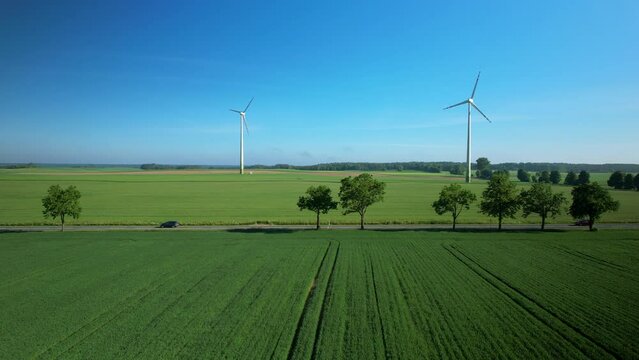 Country Road In Evergreen Fields With Towering Wind Turbines At The Background. Aerial Drone Shot