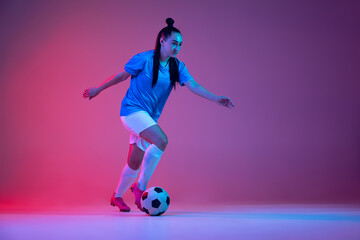 Fototapeta na wymiar Dribbling. One sportive girl, female soccer player training with football ball isolated on purple studio background in neon light. Sport, action, motion, fitness
