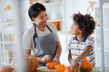 Happy young mother and her son with Afro hairstyle standing at counter and carving pumpkins together