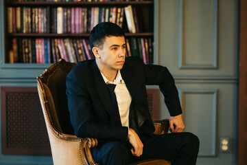 A young male manager in a suit sits in front of a bookcase on vintage armchair, leans his elbow on...
