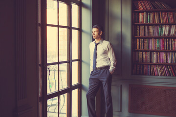 A young businessman man in trousers, white shirt and tie standing near old bookcase and looking thoughtfully to the left through panoramic window to the street
