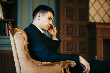 A young male manager in a suit is sitting sideways on a vintage armchair against the background of the fireplace, holding his finger at his left temple and looking thoughtfully to the right side