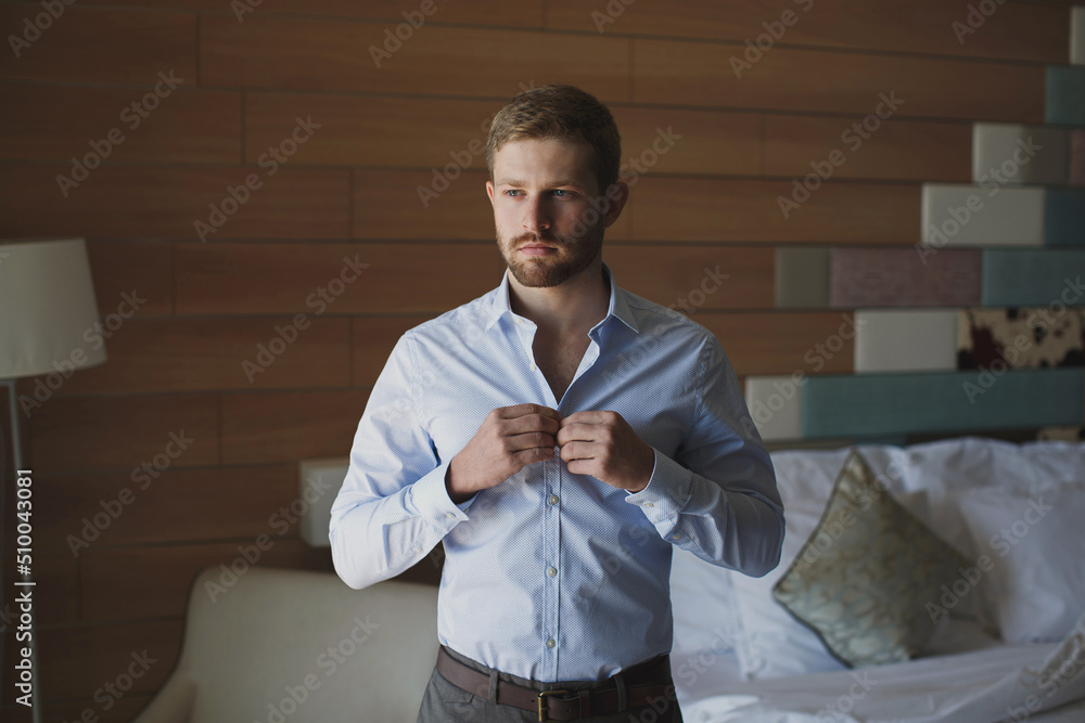 Poster handsome man groom buttoning his shirt in a hotel room