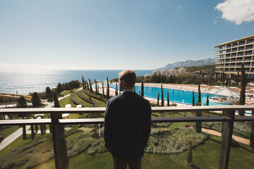 The groom stands on the balcony of the hotel and looks at the mountains and the sea