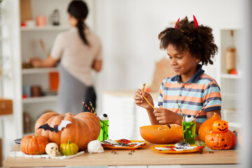 Smiling cute black boy with devils horns standing at counter with various Halloween candies and eating marmalade worm