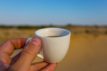 closeup human hand hold a white coffee cup among sandy desert, natural outdoor drink scene